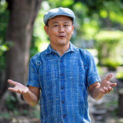 ChooChai, a tour guide, speaking with animated gestures in a green national park, wearing a blue shirt and flat cap.