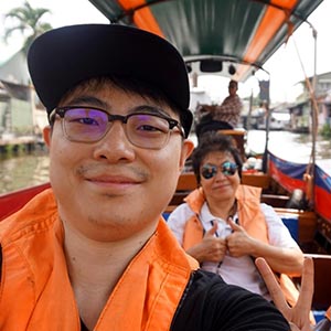 Guide Prin with Albert K. on a Bangkok canal tour, smiling aboard a long-tail boat with stilt houses and greenery in the background. 