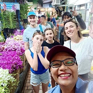 Tour guide Waza, a Thai man in a navy polo shirt, smiling beside traveler Alina G. amid vibrant floral displays at Bangkok’s flower market, surrounded by colorful bouquets and hanging garlands in a bustling, open-air stall setting.