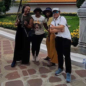 Tour guide Prin, a Thai woman in a patterned blouse, standing with traveler Amarachi O. in front of the Grand Palace in Bangkok, with intricate golden temple architecture and ornate spires shining under bright daylight.