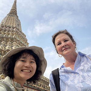Tour guide Ning, a Thai woman in a light blue blouse, smiling beside traveler Joan B. in front of the ornate spires of Wat Arun temple in Bangkok, with detailed white and gold architectural patterns in the background under clear skies.