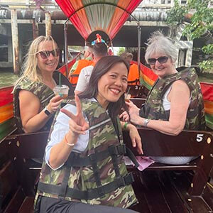 Tour guide Kanny, a Thai woman in a light top, seated beside traveler Mandy T. on a long-tail boat during a canal tour, with traditional wooden houses on stilts and lush greenery lining the waterway under partly cloudy skies.
