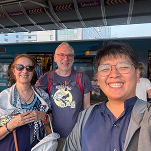 Guide Time with Neil A. at a Bangkok Skytrain station, smiling with elevated tracks and urban skyline in the background.
