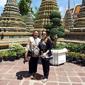 Tour guide Waza, a Thai man in a casual outfit, smiling alongside traveler Tania F. with the towering spires of Wat Arun behind them, showcasing detailed ceramic mosaics and traditional Thai temple architecture under a sunny sky.