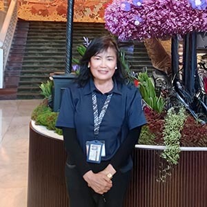 TumTum, a tour guide, smiling in a beige shirt and lanyard, standing in a hotel lobby with warm lighting and modern decor.