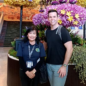 Tour guide TumTum, a Thai woman in a black top, smiling beside traveler Trent P. in a modern hotel lobby, with polished floors, warm lighting, and elegant decor in the background.”