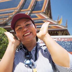 Waza, a tour guide, smiling in a light shirt and lanyard, standing in front of the Grand Palace with ornate Thai architecture behind him.