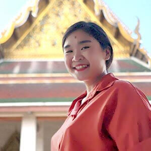 Gwen, a tour guide, smiles while leading a tour at a royal temple, wearing a pink blouse with intricate architecture visible in the background.