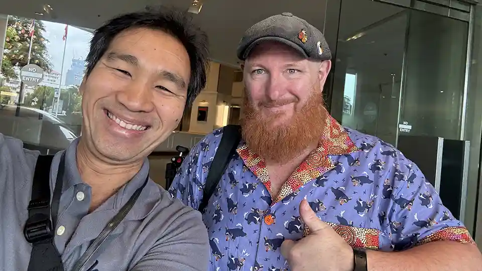 Two men are taking a selfie. The man on the left has a reddish-brown beard and is wearing a purple short-sleeved shirt with a pattern of black and white dogs, and an orange and red patterned collar. He is also wearing a gray flat cap with a small orange pin on it. He is giving a thumbs-up gesture. The man on the right has short dark hair and is wearing a gray collared shirt. Both men are smiling at the camera. In the background, there appears to be a glass entrance to a building, with a reflection of what looks like an outdoor area with some greenery and possibly text or a sign. There are also "NO ENTRY" and "ENTRY" signs visible in the reflection.