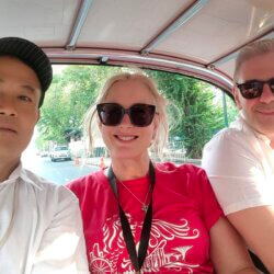 Guide ChooChai with Dave N. next to a colorful tuk-tuk in Bangkok, smiling with street life and urban scenery behind them. 